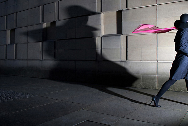 Woman in heels exiting the right of frame wearing a pink scarf and casting a distorted shadow on the wall behind her